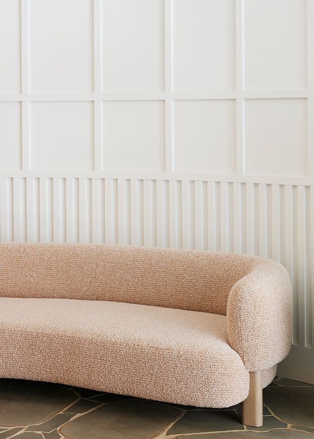 Close-up interior view of a softly upholstered, rounded sofa in a warm beige boucle-like fabric, set against a white wall with decorative rectangular panel moulding above vertical fluted panelling. The sofa stands on light timber legs over a dark, irregularly patterned stone or tile floor. The composition is minimalist, warm, and refined.