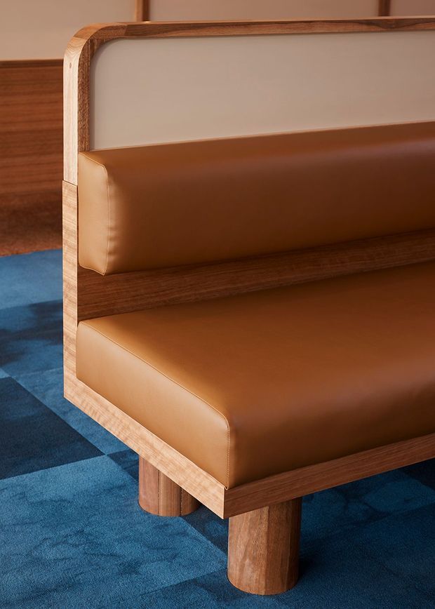 Close-up of a built-in upholstered bench or banquette with warm brown leather seat and back cushions, framed in richly grained timber with rounded edges and cylindrical timber legs. A blue patterned carpet and neutral wall panels are visible, creating a refined mid-century-inspired interior detail.