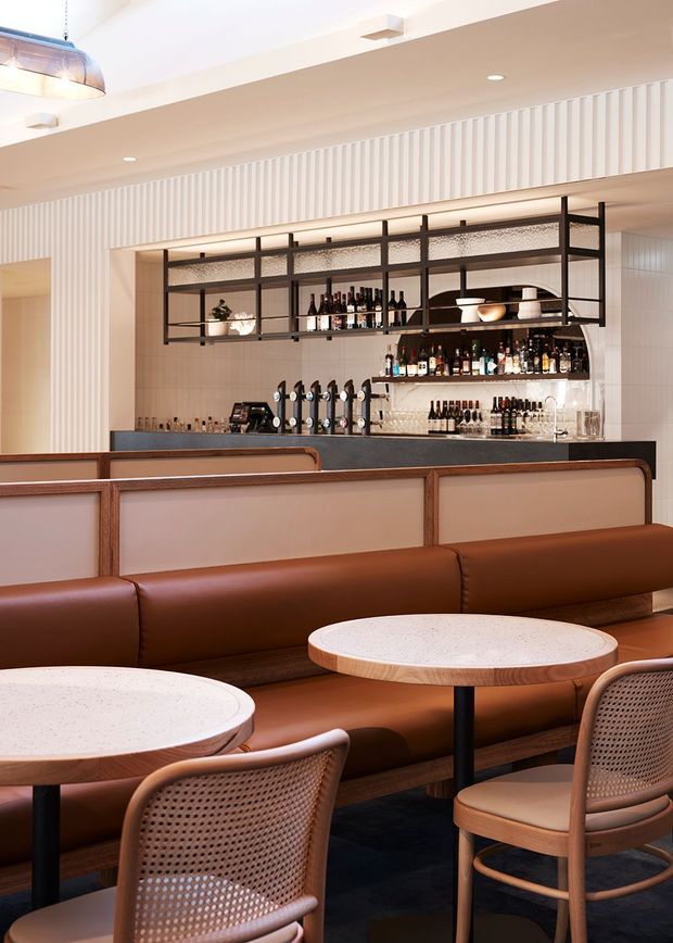 A refined contemporary bar and lounge interior with a long upholstered brown leather banquette, round café tables, cane-back chairs, and a fully stocked back bar. Open black metal shelving displays bottles and glassware, while vertical white wall panelling and warm integrated lighting create a sophisticated hospitality setting.