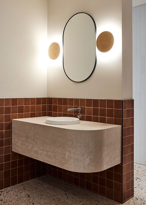 A contemporary bathroom vanity area with a floating beige concrete-look basin cabinet, white vessel sink, wall-mounted chrome tapware, and a large rounded-rectangle black-framed mirror. Warm brown square wall tiles form a half-height surround, complemented by a terrazzo-style floor and two round wall sconces.