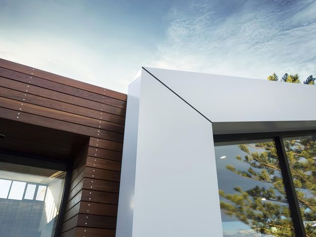 Close-up exterior architectural view showing a contemporary building facade with dark horizontal timber cladding on the left, a crisp white rendered or painted wall volume, and a large black-framed glazed opening on the right. Clean angular junctions and minimal detailing create a modern composition. Reflections of trees are visible in the glazing, with sky occupying the background.