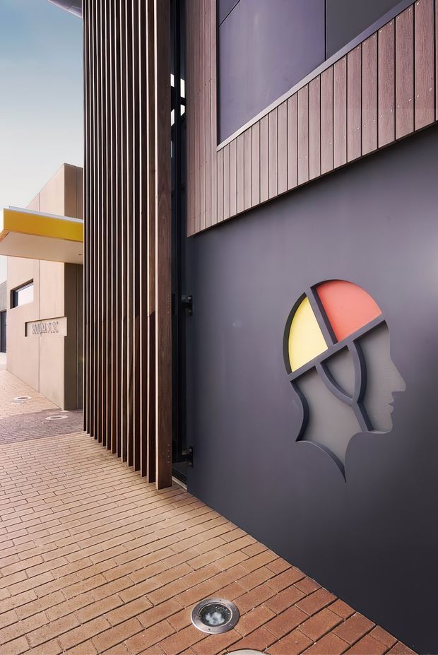 A contemporary architectural exterior passage framed by a tall vertical timber privacy screen and dark painted wall. The paved brick walkway leads past a sculptural multicoloured head-profile wall feature, with timber-look vertical cladding and a bright yellow projecting canopy visible in the background.