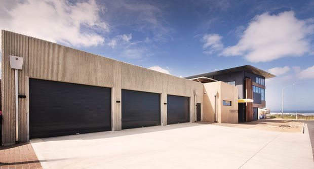 Exterior view of a contemporary low-rise commercial or industrial building with three large dark roller garage doors, a ribbed concrete or metal facade, and a smaller glazed entrance volume. A broad concrete forecourt and coastal horizon are visible to the right.