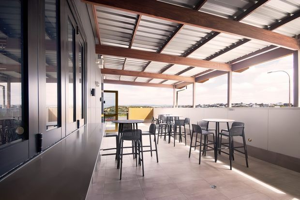 A bright, covered rooftop or elevated outdoor dining area with a corrugated metal roof supported by exposed dark timber or metal framing. The space has pale tiled flooring, a long enclosed wall with glazing on one side, high communal tables and bar-height seating, and open sides overlooking a low-density urban or coastal landscape. The setting appears contemporary and functional, suitable for hospitality or shared amenity use.