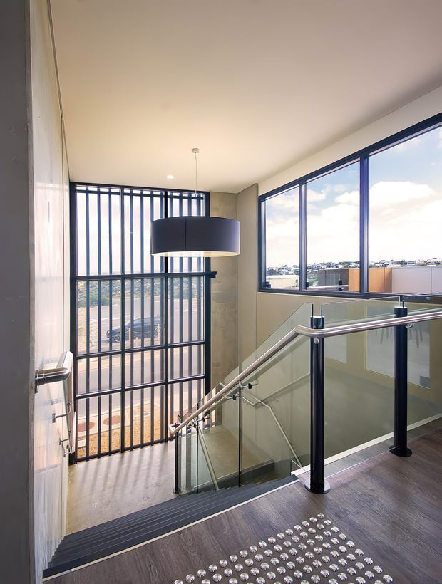 A bright, modern stairway and landing with dark timber-look flooring, a glass balustrade with stainless-steel handrails, and black metal security-style vertical bars at the glazed entry. Large blue-framed windows overlook a suburban or urban landscape. A large black drum pendant hangs above the stairwell, while tactile silver floor markers provide contrast on the landing.