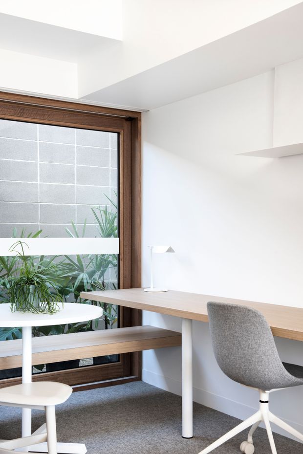 Bright, minimalist home office nook with a long light-wood desk spanning a white wall, a grey upholstered task chair, white desk lamps, and a built-in window bench. The large timber-framed window brings in natural light and looks onto a planted light-grey masonry courtyard wall. Grey carpet, crisp white walls, and simple integrated shelving create a clean contemporary workspace.