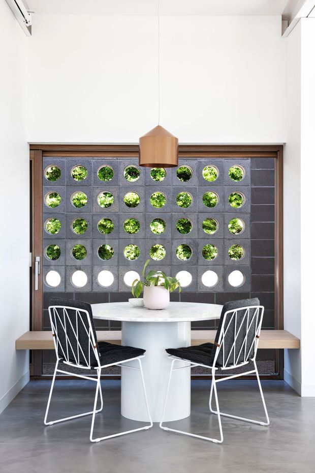 Bright, compact dining area with a round white table and sculptural pedestal base, two black-and-white wire-frame chairs, and a timber pendant light. The space is set against a distinctive dark concrete breeze-block screen with circular openings revealing greenery beyond, framed by dark timber joinery. A built-in timber bench runs along the wall, and the polished grey floor and white walls create a clean contemporary setting.