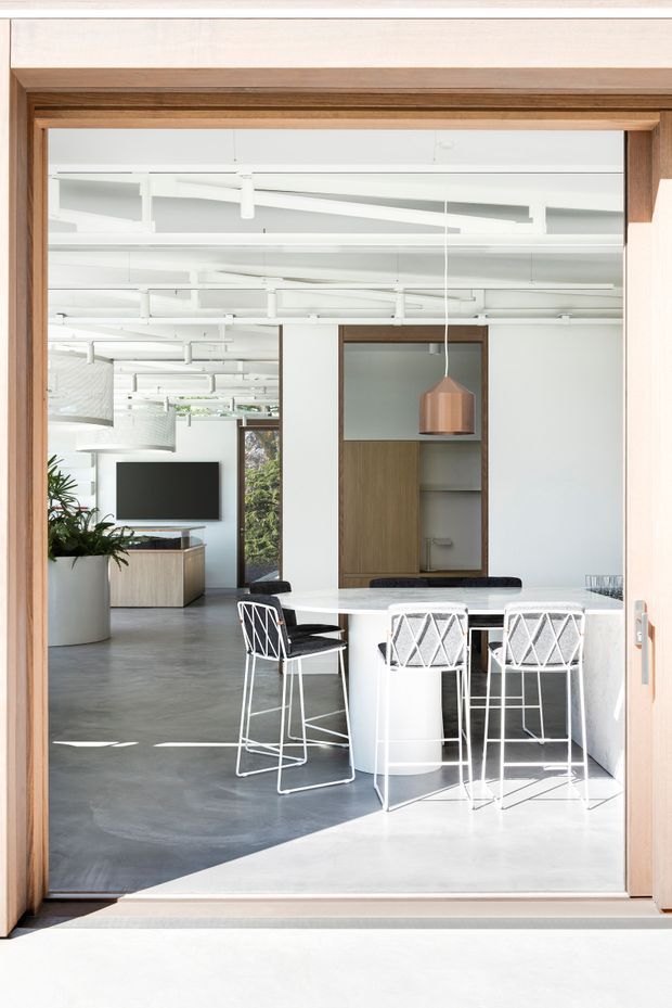 Bright contemporary open-plan interior viewed through a timber-framed glazed doorway. A white island or bar table with white cylindrical base and wire-frame bar stools occupies the foreground, while a dining/work area, built-in timber shelving, copper pendant light, television, indoor plants and polished grey concrete flooring extend into the background. The ceiling is white with exposed services and linear suspended lighting, and strong daylight creates a clean, airy atmosphere.