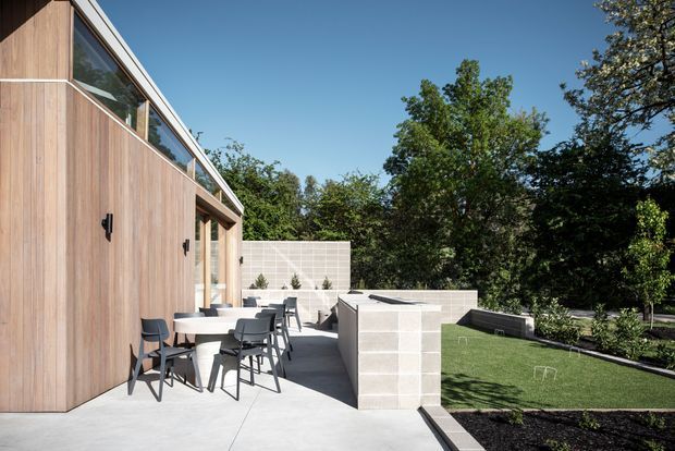 Contemporary outdoor terrace beside a timber-clad building, featuring a dining table with chairs, pale concrete paving, rendered masonry privacy walls, raised planters, and a neatly landscaped lawn and garden. Large windows and clean horizontal lines contribute to a modern architectural composition.