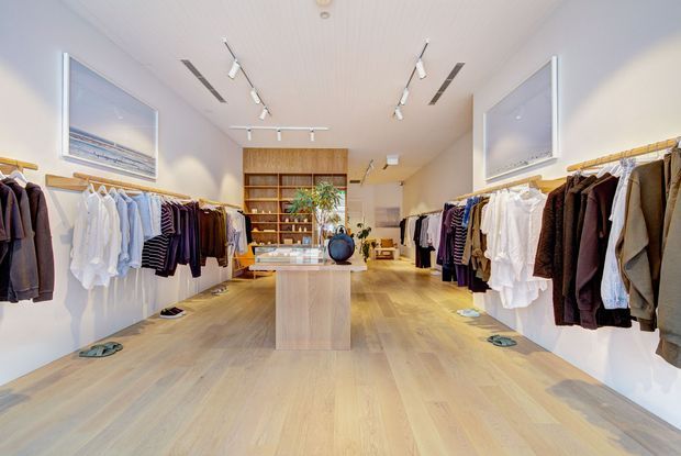 Bright, contemporary clothing boutique with a spacious open retail floor, light timber flooring, white walls, wall-mounted clothing rails, central timber display island, rear shelving, and track lighting. Framed artwork and neatly arranged garments contribute to a clean, refined retail interior.