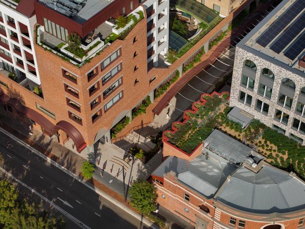 Aerial view of a dense urban architectural precinct featuring a prominent multi-storey red-brick building with rooftop terraces and planted roof gardens, adjacent to a light stone-clad building with rooftop solar panels. Landscaped podiums, pedestrian paths, a covered parking area, and a curved heritage-style brick building are visible around the street edge.