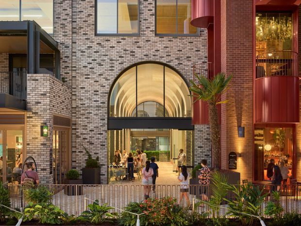 A contemporary multi-storey commercial or mixed-use building entrance viewed from an outdoor landscaped area. The facade combines patterned brickwork, dark metal framing, and bold red architectural elements. A large arched glazed entrance canopy and tall windows reveal a bright interior with people and dining or retail activity. Tropical planting and metal railings occupy the foreground.