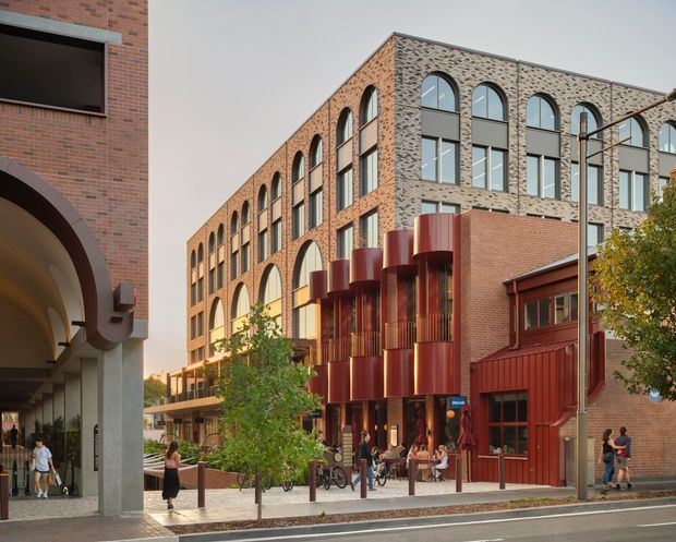 Street-level exterior view of a contemporary multi-storey brick building with a long facade of repeated arched windows, contrasting grey masonry, and a prominent sculptural red-metal entrance volume. The building fronts a paved pedestrian area and roadway, with adjoining brick architecture visible at the left.