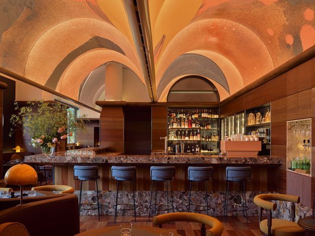 A richly detailed, intimate cocktail bar interior with a long stone-topped bar, dark timber cabinetry, illuminated bottle shelving, glassware displays, and upholstered bar stools. The space is defined by an ornate vaulted ceiling with sculptural ribbed arches in warm terracotta, orange, and bronze tones, creating a dramatic and luxurious atmosphere.