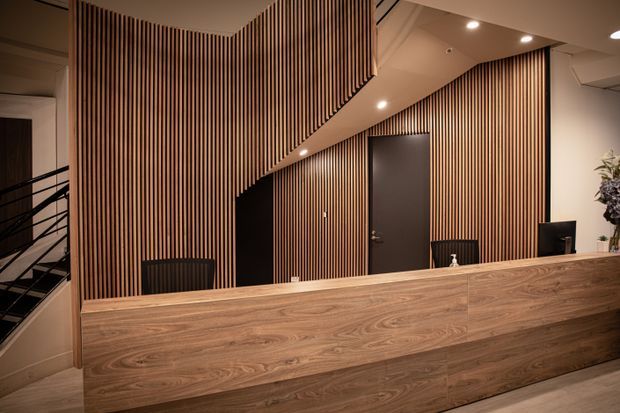 A warmly lit contemporary reception desk with extensive vertical timber slat wall panelling, a large wood-grain front counter, dark charcoal door and cabinetry, recessed ceiling lights, and a stairway visible at the left. The space has a refined commercial lobby or reception feel.