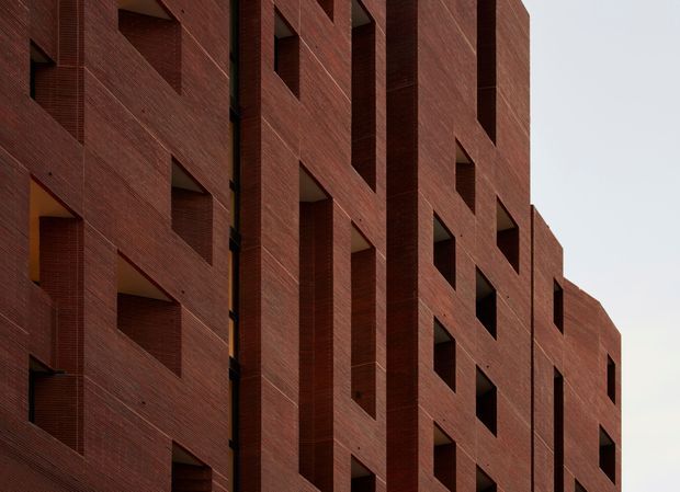 Close-up exterior view of a multi-storey building with a strongly geometric facade. Deeply recessed rectangular window openings and projecting vertical brick fins create a rhythmic, sculptural composition. The facade is clad in dark red-brown textured brick, with minimal visible glazing and an overcast sky.