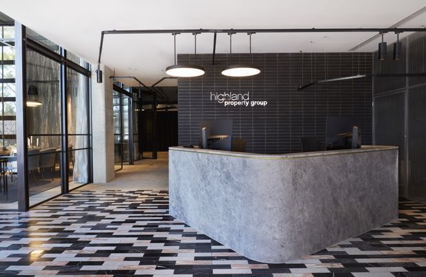 A contemporary commercial reception area with a large curved grey stone or concrete reception desk, dark tiled feature wall, exposed black ceiling services, and black-framed glass partitions. Geometric patterned floor tiles and suspended circular pendant lights create a refined industrial office-lobby aesthetic. The Highland Property Group logo is displayed behind the reception desk.