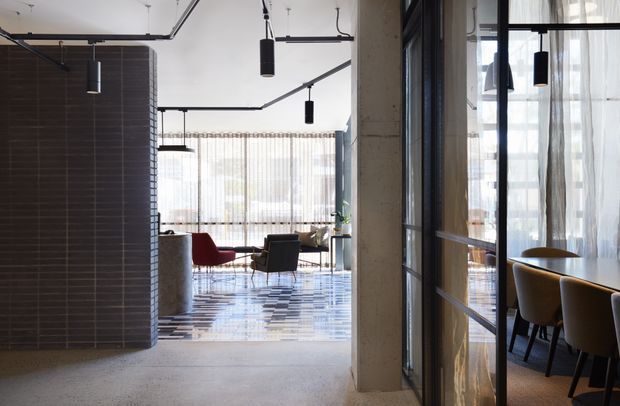 A refined contemporary commercial interior with a polished patterned floor, dark tiled cylindrical partition, exposed black linear services and suspended cylindrical pendant lights. The open lounge area contains modern seating near full-height sheer curtains, while a glazed dining or meeting room with a timber table and upholstered chairs is visible on the right.