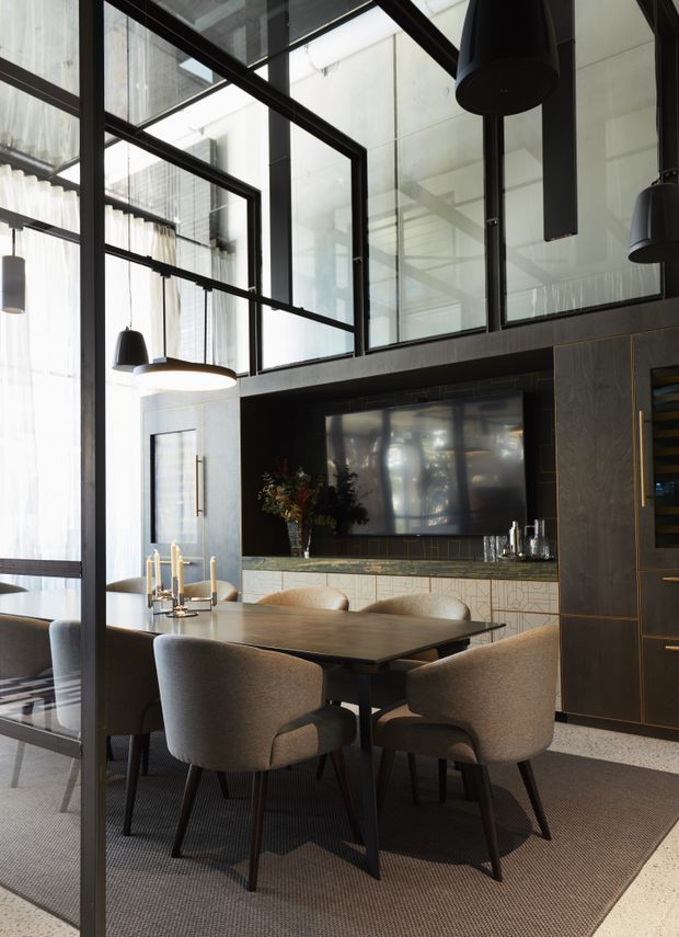 A refined dining area with a dark rectangular dining table and upholstered chairs, enclosed by black-framed internal glazing. A large wall-mounted television, integrated cabinetry, decorative lighting, and a drinks cabinet create a sophisticated open-plan entertaining space. The room has high ceilings and abundant filtered daylight.