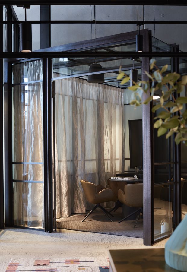 A moody contemporary interior viewed through black steel-framed glazing and doors. The space contains a small dining or meeting table with upholstered brown armchairs, sheer floor-to-ceiling curtains, exposed structural steel beams, pendant lighting, and polished concrete or terrazzo flooring. A leafy plant and partial furniture appear in the foreground.