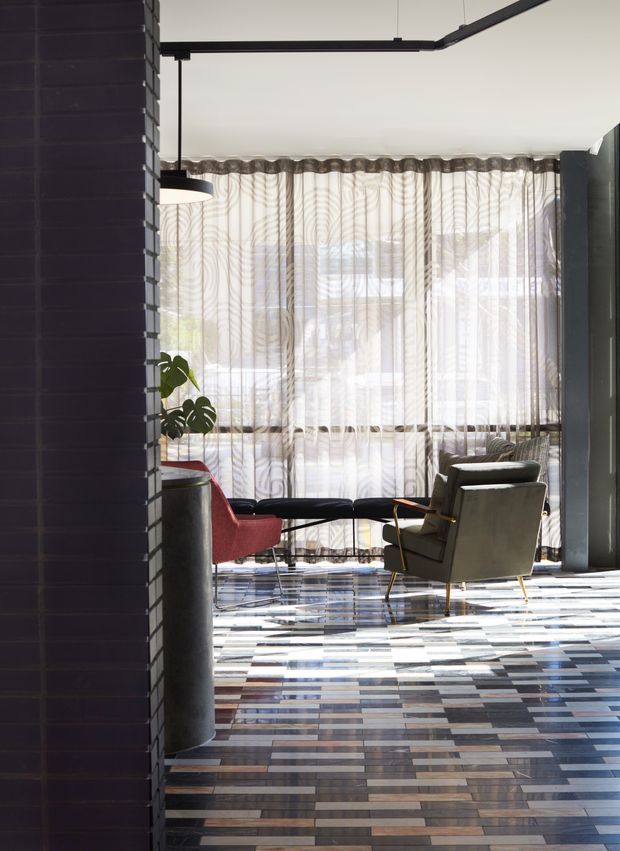 A stylish, light-filled living room with floor-to-ceiling sheer patterned curtains, a dark tiled or brick feature wall, exposed black ceiling-mounted fittings, and a striking patchwork timber-look floor. Two lounge chairs and a low dark sofa form a seating area near the windows, with a tall indoor plant and a cylindrical side element adding visual interest. The space has a refined mid-century-inspired character with strong material and colour contrast.