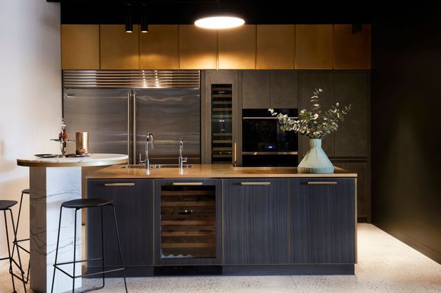 A sophisticated contemporary kitchen with a large central island, integrated dark cabinetry, built-in wine storage, stainless-steel refrigeration, and a compact breakfast bar. Warm bronze-toned upper cabinetry contrasts with the dark vertical-grain joinery, while pendant lighting and a pale terrazzo-style floor provide balance.