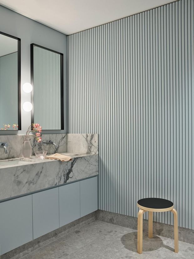 A refined contemporary bathroom featuring a long floating vanity in pale blue-grey cabinetry, a dramatic grey-veined marble benchtop and backsplash, black-framed mirrored cabinets, and a vertically fluted blue-grey feature wall. Grey stone flooring and skirting, spherical mirror lighting, and a simple timber-and-black stool complete the calm, modern scheme.