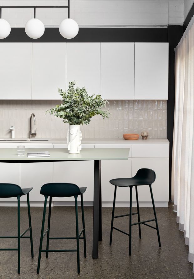 A bright, refined contemporary kitchen with a long pale island and dark sculptural bar stools. The room features flat-panel white cabinetry, a glossy light tiled splashback, a dark polished-looking floor, black-framed spherical pendant lights, and sheer floor-to-ceiling curtains. A stainless-steel tap and sink sit beneath the wall cabinets, while a vase of greenery provides a natural focal point.