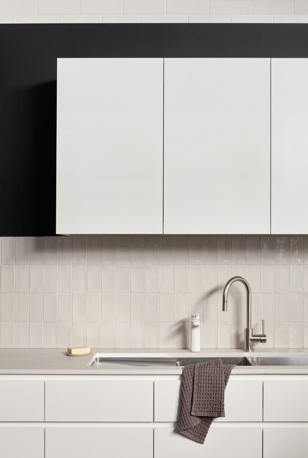 A minimalist contemporary kitchen focused on a clean white and grey fit-out. Flat-front white overhead and lower cabinetry sits against a matte black upper wall and pale vertically gridded ceramic tile splashback. A light grey benchtop incorporates a stainless-steel sink and tall curved chrome tap, with a neutral textured tea towel adding a subtle soft accent.