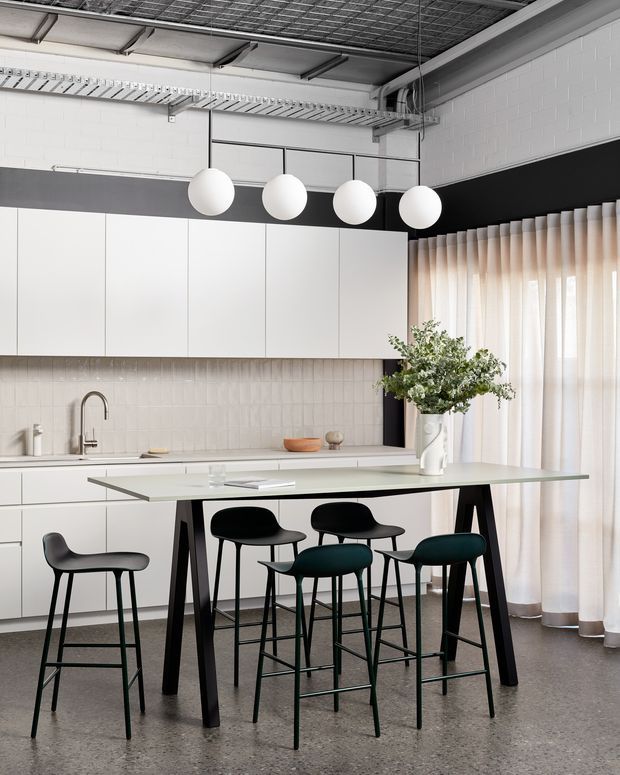 A bright contemporary kitchen with extensive white flat-panel cabinetry, a pale tiled splashback, and a long island with a thin light benchtop supported by dark angled legs. Four spherical pendant lights hang above the island, which is surrounded by dark bar stools. The room has an exposed industrial-style ceiling with visible services, white painted brick walls, black horizontal wall banding, polished grey flooring, and full-height sheer curtains.