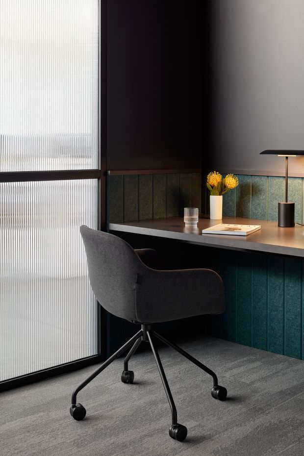 A compact, refined home office nook with a dark built-in desk and matching teal-green vertical wall panelling. A charcoal upholstered swivel office chair sits on a grey carpeted floor, beside a large ribbed translucent window or screen in a dark frame. The desk includes a slim black table lamp, a small vase of yellow flowers, a glass, and books. The atmosphere is moody, sophisticated, and contemporary.