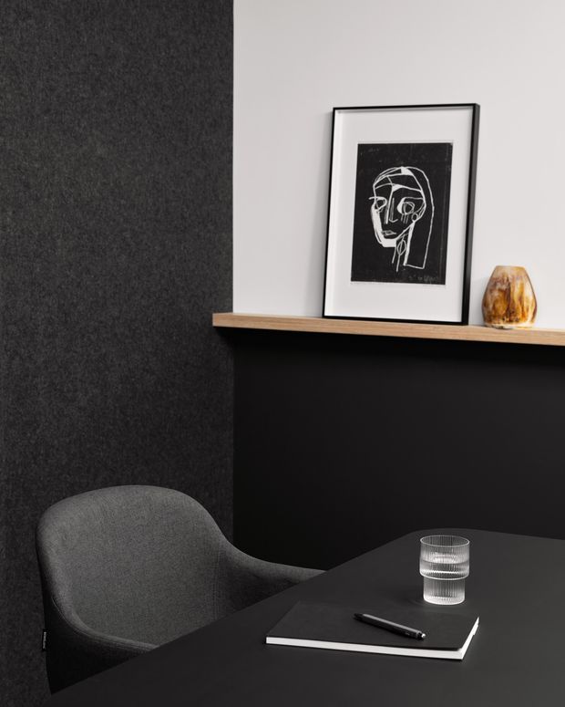 A refined, dark-toned interior vignette featuring a charcoal upholstered armchair beside a black desk or table. A black feature wall with a narrow light-wood shelf supports framed monochrome artwork and a small amber-toned decorative object. A notebook, pen, and ribbed drinking glass sit on the tabletop, creating a minimalist workspace or study setting.