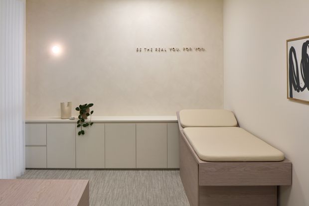 A softly lit, minimalist treatment room with a pale upholstered treatment bed, built-in low cabinetry along the back wall, a neutral textured carpet, sheer curtains, and understated wall art. The space has warm beige and light timber tones with clean, uncluttered lines.