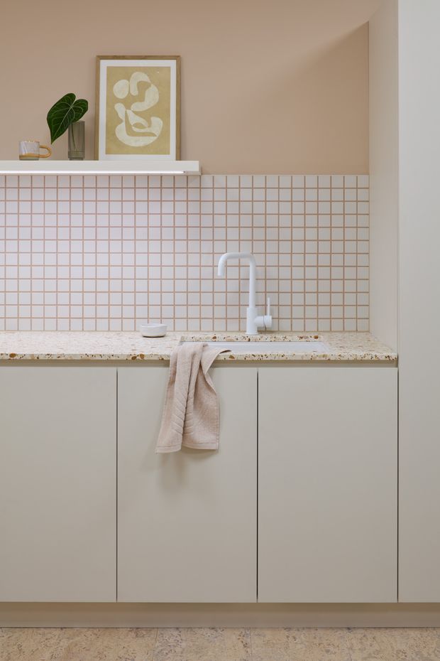 A compact, softly styled kitchen or kitchenette with pale beige flat-panel cabinetry, a speckled light stone benchtop, and a backsplash of small square blush-beige tiles with contrasting grout. A white gooseneck mixer tap and integrated sink sit beneath the tiled wall. A floating white shelf displays a framed abstract artwork, a potted plant, and a small glass, while a neutral linen tea towel adds warmth. The space has a calm, minimalist contemporary aesthetic with warm neutral tones.