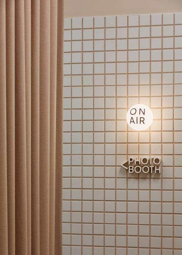 A close-up of a softly lit commercial interior wall featuring a grid of small square pale tiles with warm grout. A full-height beige curtain occupies the left side, while illuminated wall signage reads “ON AIR” and “PHOTO BOOTH,” indicating a photo booth or studio setting. The composition is minimalist and warm, with strong geometric repetition and concealed or subtle lighting.