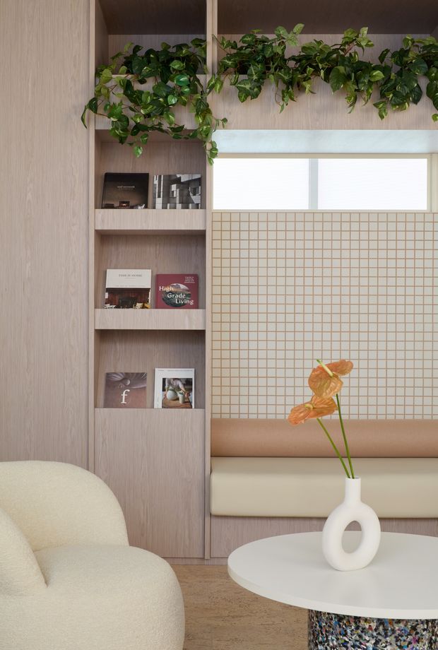 A refined contemporary interior vignette featuring light timber built-in shelving and cabinetry, trailing green plants, curated books, a large tiled feature wall with a horizontal window, and integrated upholstered seating. A rounded cream boucle armchair and sculptural white vase sit beside a circular coffee table with a terrazzo-like recycled aggregate base. The palette is warm and neutral with soft natural textures and restrained organic decoration.