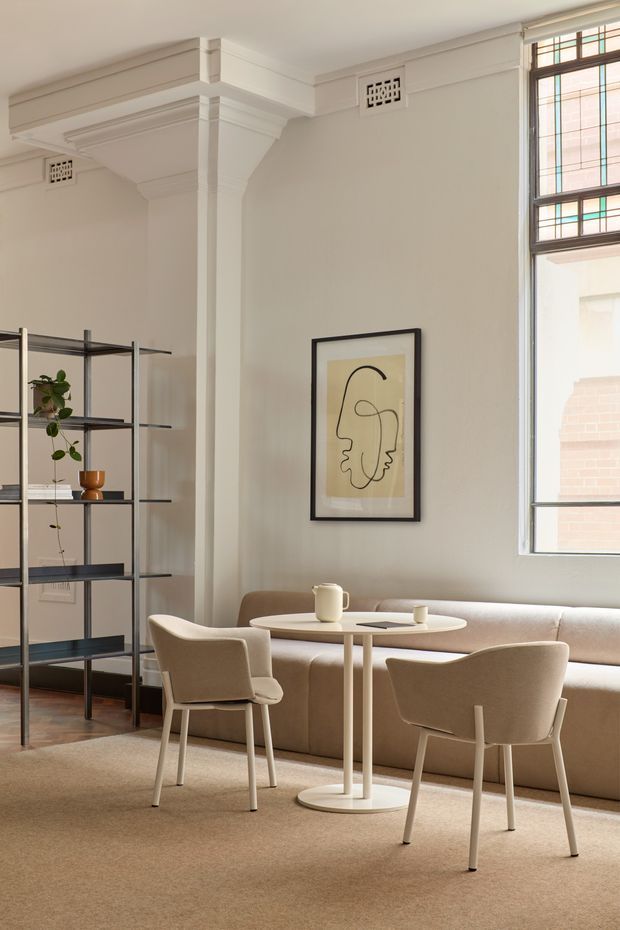 A refined sitting and dining area in a bright heritage-style interior. The room features ornate painted wall and ceiling mouldings, a tall stained-glass window, warm neutral walls, a large upholstered banquette, a small round pedestal table, two upholstered dining chairs, a black metal open shelving unit, and minimalist framed line artwork. Timber flooring is partly covered by a large beige carpet.