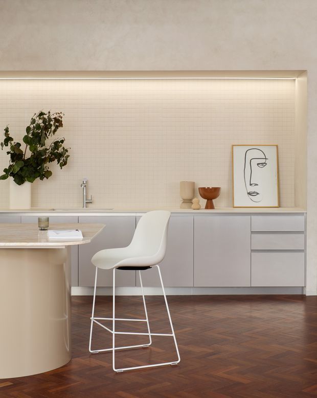 A refined contemporary kitchen with a pale, minimalist palette. Flat-front light grey cabinetry runs along the rear wall beneath a softly illuminated cream tiled backsplash, with a slim benchtop and integrated sink and tap. A curved island with a rounded beige-toned base occupies the foreground, paired with a white sculptural bar stool. Dark herringbone timber flooring adds warmth, while a potted plant, ceramic vessels and framed line art provide restrained decorative accents.
