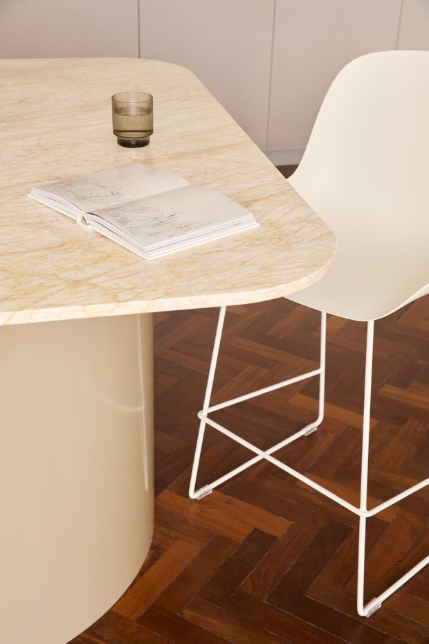 Close-up interior view of a refined dining or kitchen island setting. A rounded rectangular light stone or marble-like tabletop with subtle veining sits on a curved beige pedestal base, paired with a white molded bar stool. The space features dark warm timber parquet flooring, pale walls, and a restrained contemporary aesthetic. A glass and open book add minimal styling.