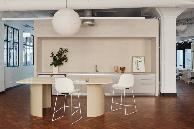 A bright open-plan commercial-style kitchen and dining area with a long pale island bench, integrated light cabinetry, two white bar stools, and a large spherical pendant light. The space has exposed white services and ducts, large black-framed windows, white structural columns, and a dark herringbone timber floor. A small adjoining workspace or seating area is visible through the opening on the right.