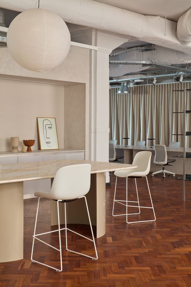A refined contemporary commercial interior combining a kitchenette or hospitality counter with an open office meeting area. The space features a pale stone-look island with white bar stools, light built-in cabinetry, a sculptural paper pendant, warm herringbone timber flooring, vertical acoustic curtains or screens, exposed services painted white, and soft neutral finishes. Workstations and task lighting are visible in the background.