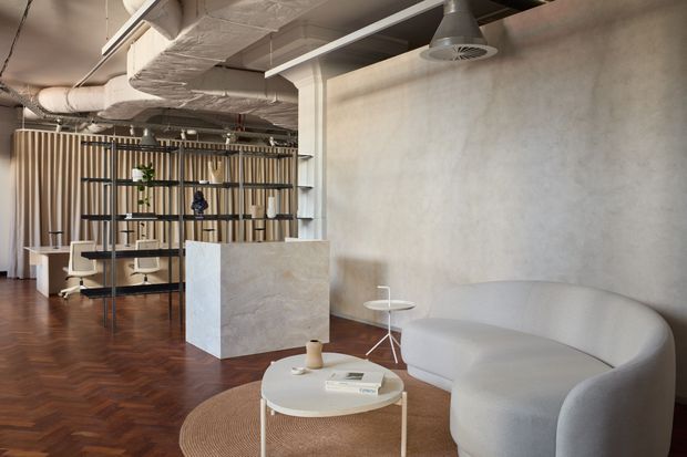 Contemporary open-plan commercial interior with a lounge area in the foreground and workstations behind a sculptural shelving divider. The space features exposed services and ductwork, a textured pale plaster wall, warm herringbone timber flooring, vertical timber slat screening, curved upholstered seating, and minimalist round tables. The palette is warm neutral with white, cream, light timber, and brown tones.