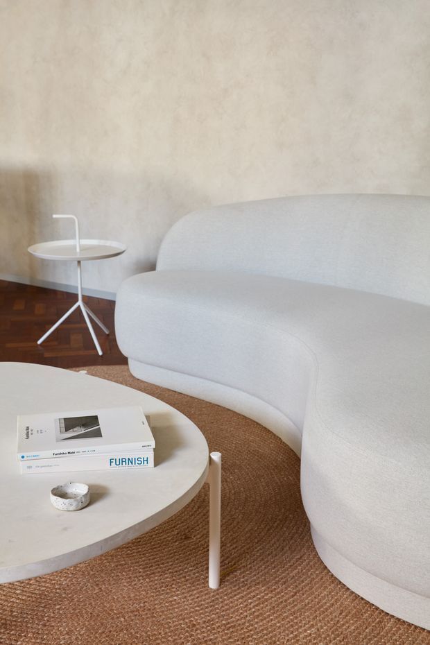 A softly styled living room vignette featuring a large curved off-white upholstered sofa, a rounded pale stone or concrete coffee table, and a slim white tripod side table. A textured natural-fibre rug covers much of the floor, with dark parquet timber flooring visible near the wall. The warm plaster-textured wall and restrained furnishings create a calm, minimalist interior.