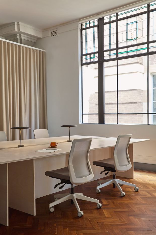 Bright, refined office interior with a long pale built-in worktable, ergonomic white office chairs, slim task lamps, and a dramatic large leaded-glass window with geometric detailing. Cream drapery, white walls and trim, and a warm herringbone parquet floor create a calm contemporary workspace with historic architectural character.