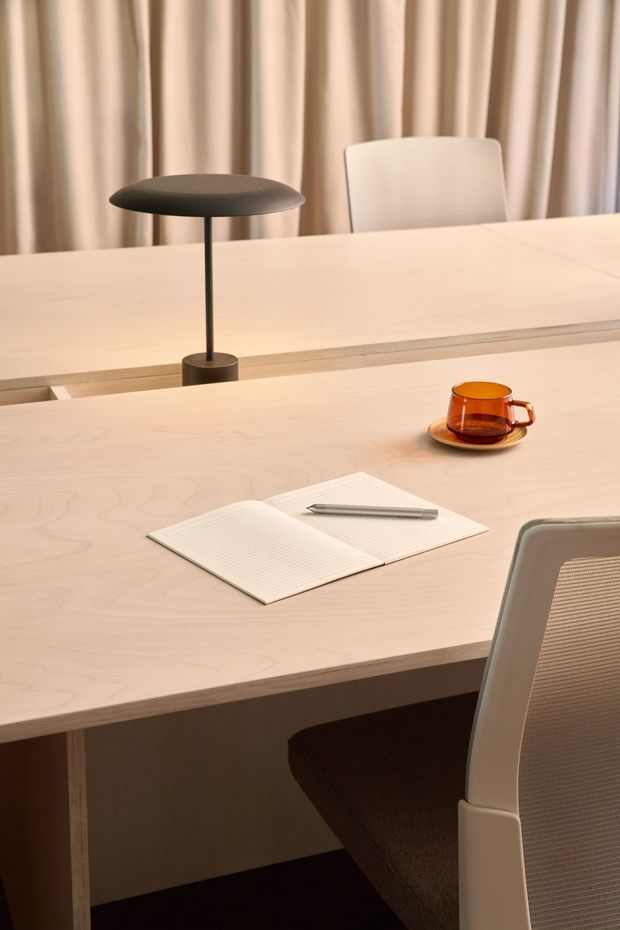 A softly lit contemporary workspace or dining area featuring a large pale timber table, a slim black desk lamp, an amber glass cup and saucer, an open notebook with pen, and modern upholstered chairs. Full-height beige curtains create a warm, calm backdrop.