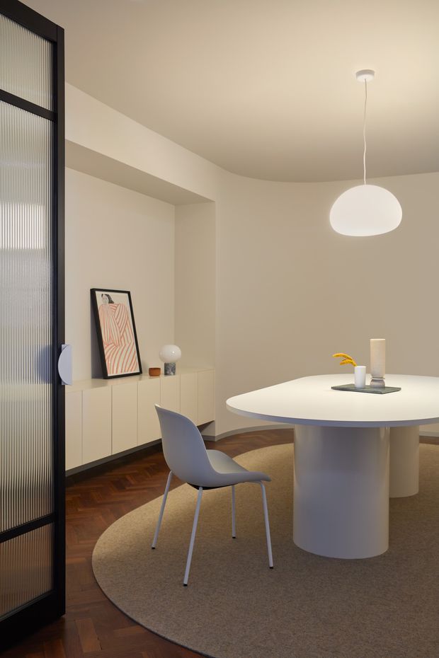 A refined contemporary dining area with a large round white pedestal table, sculptural white pendant light, curved pale dining chair, and low built-in cabinetry. Warm herringbone timber flooring is partially covered by a large neutral round rug. A black-framed fluted-glass door or partition defines the left edge, while soft beige walls and minimalist artwork create a calm, modern interior.