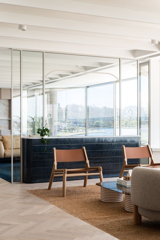 Bright contemporary living room with floor-to-ceiling glazing overlooking a waterfront urban landscape. The room features a pale herringbone timber floor, a dark ribbed built-in divider or joinery element, warm timber-framed lounge chairs with leather or tan upholstery, a textured neutral sofa, a natural woven rug, and a sculptural round coffee table. White exposed-beam ceilings and extensive internal glass partitions create an airy, open-plan feel.
