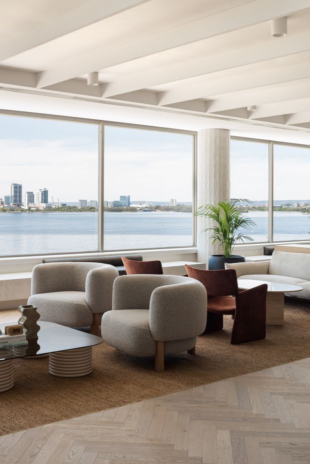 A bright, contemporary waterfront living room with expansive floor-to-ceiling windows overlooking a bay and distant city skyline. The room features sculptural upholstered lounge chairs, a white sofa, dark wood accent chairs, a rounded coffee table, a large textured rug, a potted palm, and pale timber parquet flooring. Exposed white ceiling beams and cylindrical downlights add architectural character.