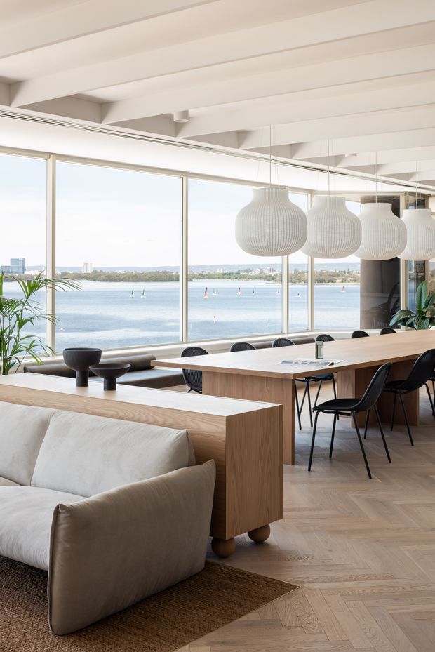 An airy open-plan waterfront interior combining a living area and dining space. The room features extensive floor-to-ceiling glazing overlooking the water, white exposed ceiling beams, pale timber parquet flooring, a large light timber dining table and integrated console, sculptural white pendant lights, black dining chairs, and a soft neutral sofa with a textured rug. The overall design is refined, contemporary, and warm with abundant natural light.