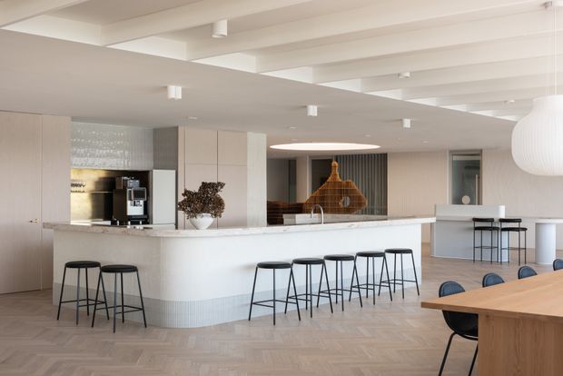 A bright, spacious open-plan kitchen and dining area with a large white island, integrated cabinetry, coffee station, and extensive bar seating. The room features pale timber herringbone flooring, white structural ceiling beams, ribbed island detailing, a sculptural pendant light, and a timber dining table in the foreground. Neutral finishes and clean-lined furnishings create a refined contemporary interior.